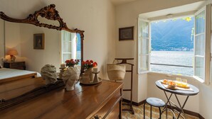 Ein Zimmer mit einem hölzernen Schminktisch, einem Spiegel, einem Tisch mit Frühstücksgebäck und -zutaten, Blick auf einen See und einem Fenster mit Bergblick.