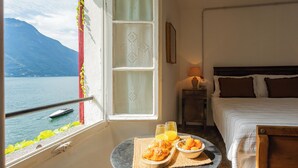 Ein Hotelzimmer mit Bett, einem kleinen Tisch mit Frühstück, einem Fenster mit Blick auf einen See und Berge sowie einem Nachttisch mit Lampe.