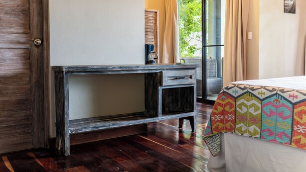 Une pièce avec un bureau en bois, un lit recouvert d’une couverture aux couleurs vives, et une vue sur la verdure à l’extérieur.
