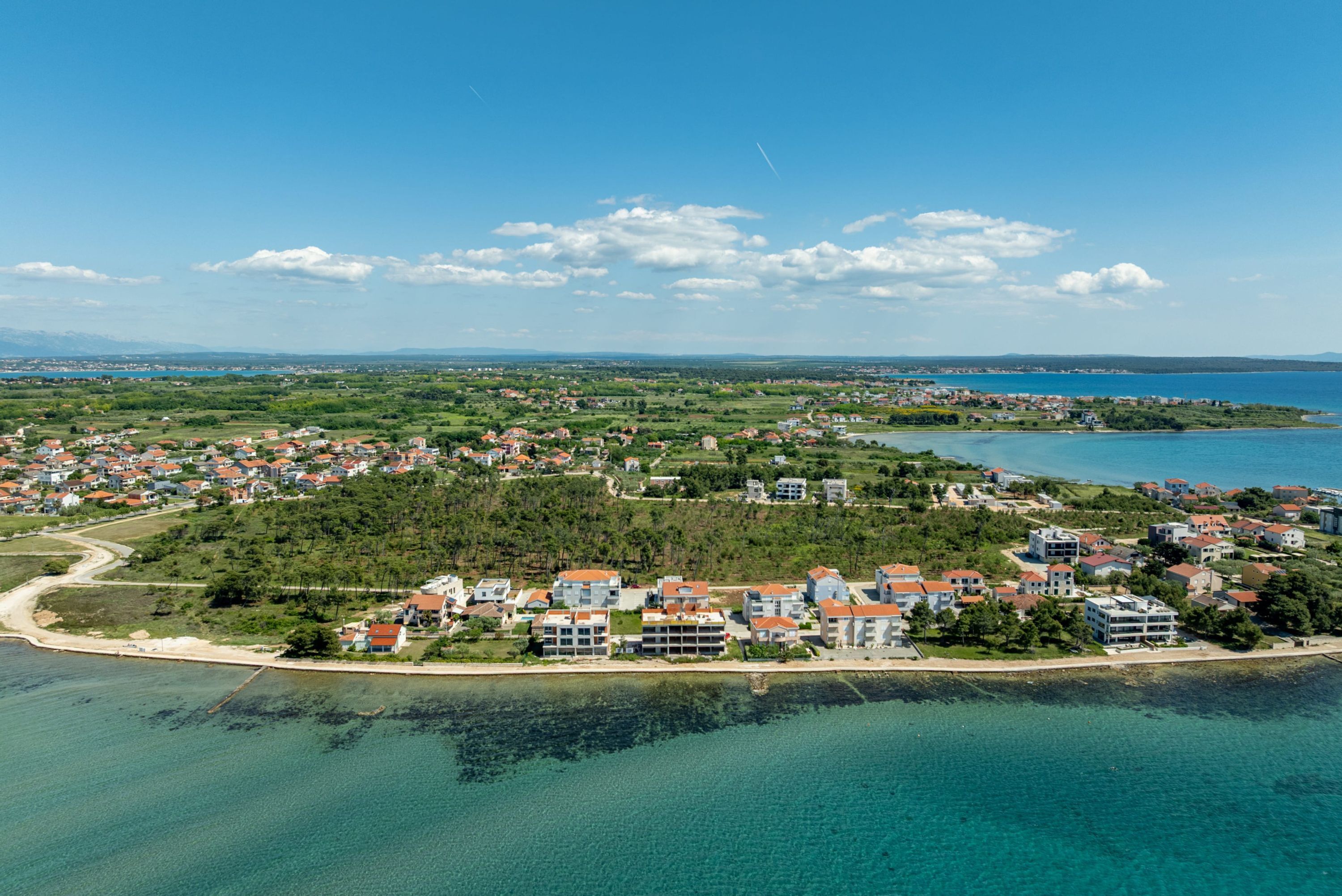 Luchtbeeld van een kustplaatsje met woongebouwen, een strand en helderblauw water.