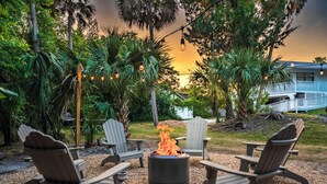 A fire pit with four Adirondack chairs in a garden setting with a view of a house in the background.