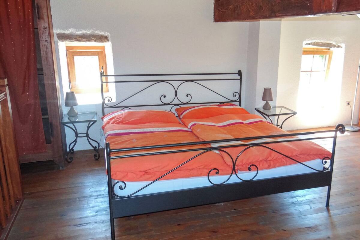 A metal-framed bed with a red and white bedspread, a bedside table with a lamp, and a wooden bunk bed in the background.
