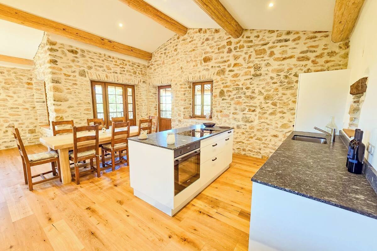 A rustic kitchen with stone walls, wooden beams, a central island, and a dining area with wooden chairs and a table.