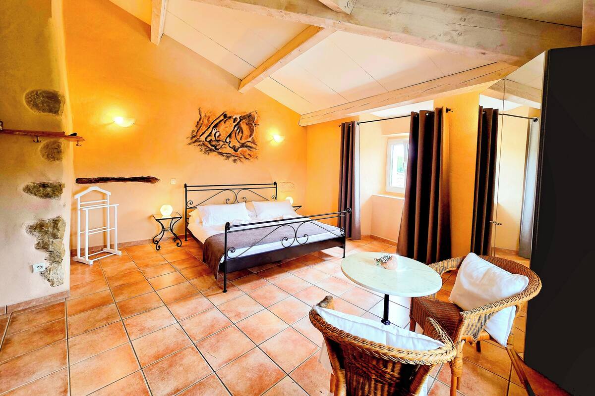 A bedroom with a bed, a small table, wicker chairs, a window, and a wall-mounted artwork.