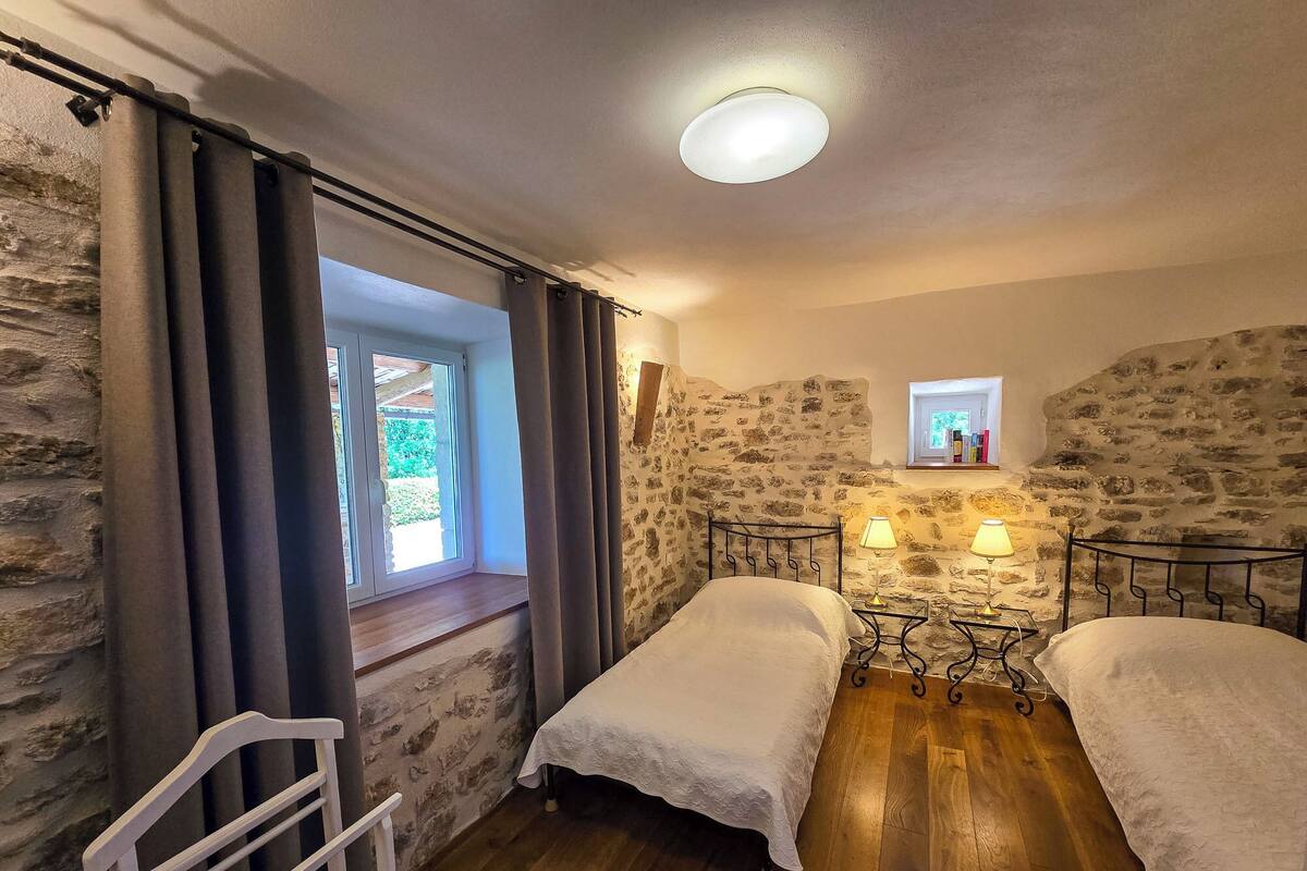 A bedroom with two single beds, a window with curtains, a stone wall, a small table with lamps, and a ceiling light.