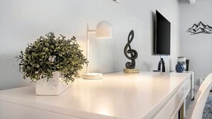 A modern, minimalist room with a white table, a potted plant, a desk lamp, a decorative treble clef, and a wall-mounted TV. - Beautiful remodeled 1-bedroom condo in Littleton/ Englewood with Pool (Englewood)