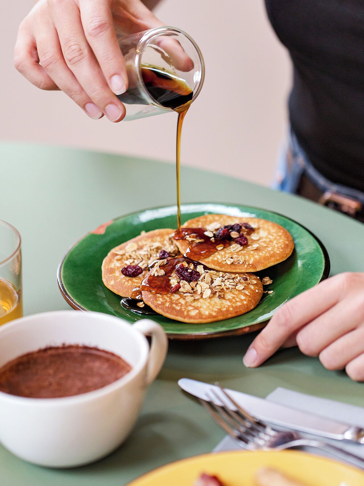 A person pouring syrup over pancakes on a green plate, with a cup of coffee and utensils on the table.