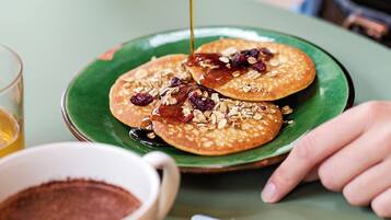 A person pouring syrup over pancakes on a green plate, with a cup of coffee and utensils on the table.
