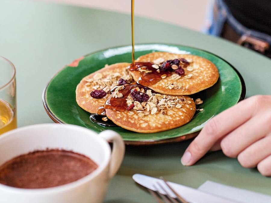 Iemand giet siroop over pannenkoeken op een groen bord, met een kop koffie en bestek op tafel.