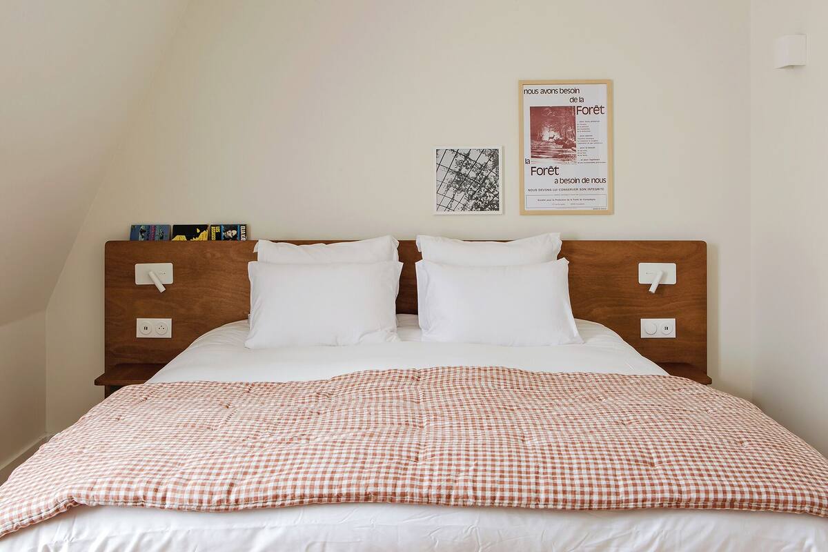 Ein ordentlich bezogenes Bett mit einer karierten Decke, zwei weißen Kissen und einem hölzernen Kopfteil mit Nachttischen. An der Wand hängen zwei gerahmte Poster, eines mit Text und das andere mit einem Schwarz-Weiß-Bild.