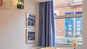 A dining area with a wooden table, white chairs with blue cushions, framed photographs on the wall, and a window with a view of a building. - Alfred Hotels Toulouse Centre Wilson - Hôtel Rénové  (Toulouse)
