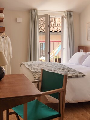 A hotel room with a bed, a desk, a chair, a window with curtains, and a view of a building with a tiled roof.