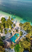 Vista aérea de un complejo turístico en la playa con piscina, palmeras y embarcaciones en el agua.