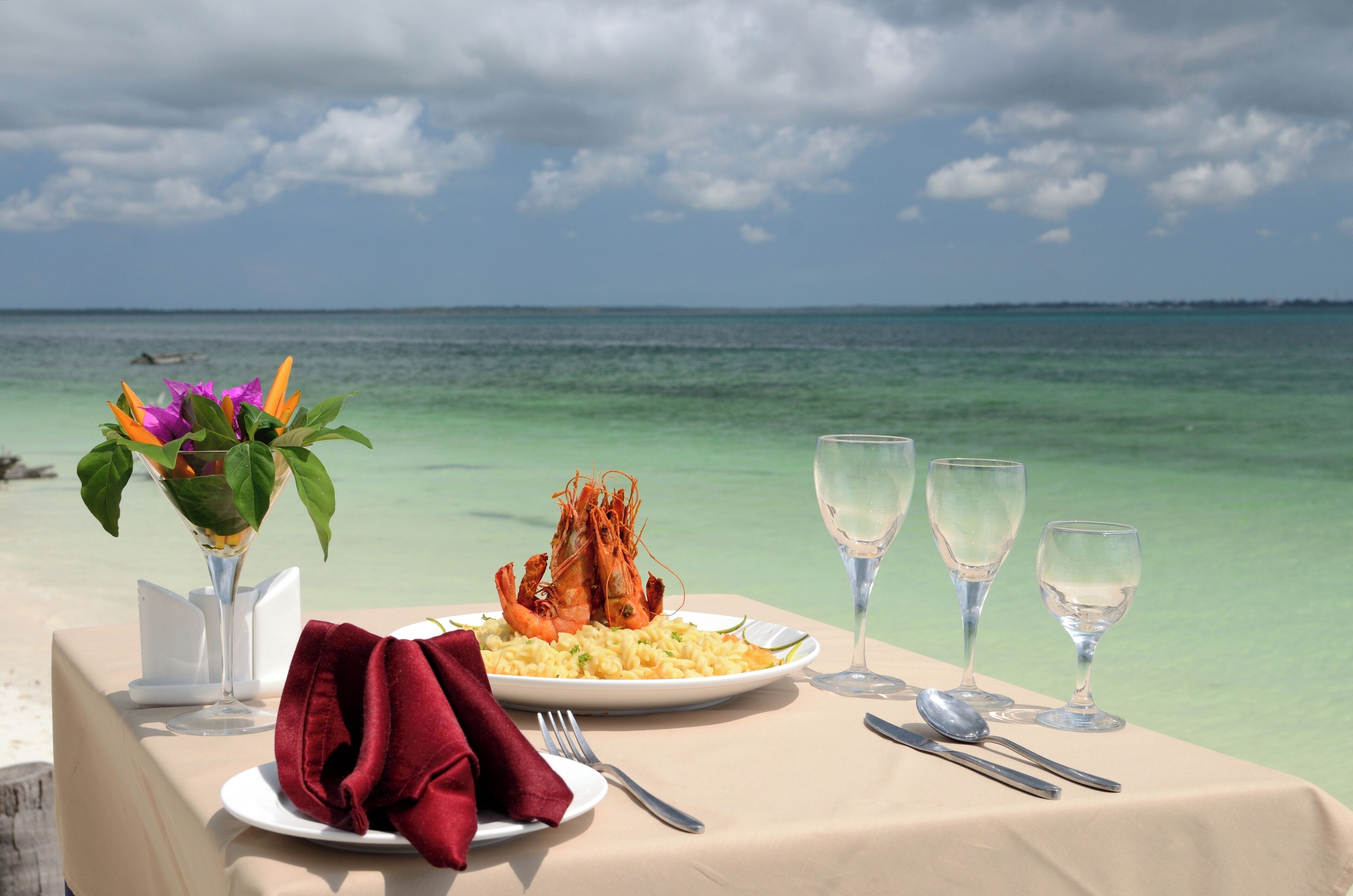 A table set with a plate of food, a glass of water, and a red napkin on a beach with a view of the ocean.