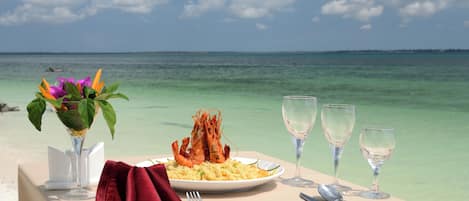 A table set with a plate of food, a glass of water, and a red napkin on a beach with a view of the ocean.