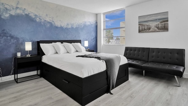 A modern bedroom with a large bed, a black sofa, and a city view through the window. - Ocean Dr 1BR Miami Beach Getaway (Miami Beach)