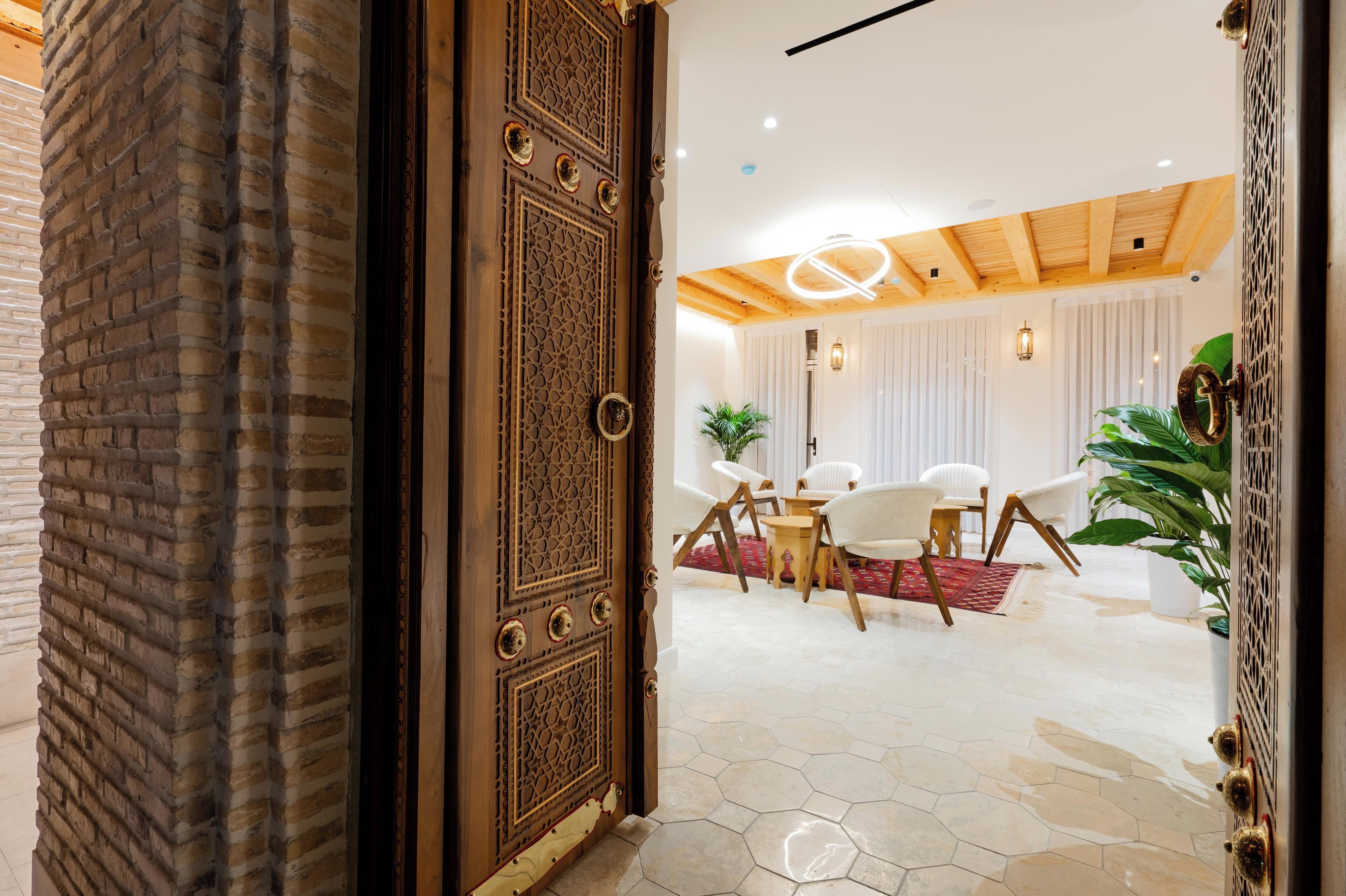 A room with a brick wall, a wooden door with intricate carvings, a round ceiling light, and a dining area with white chairs and a small table.