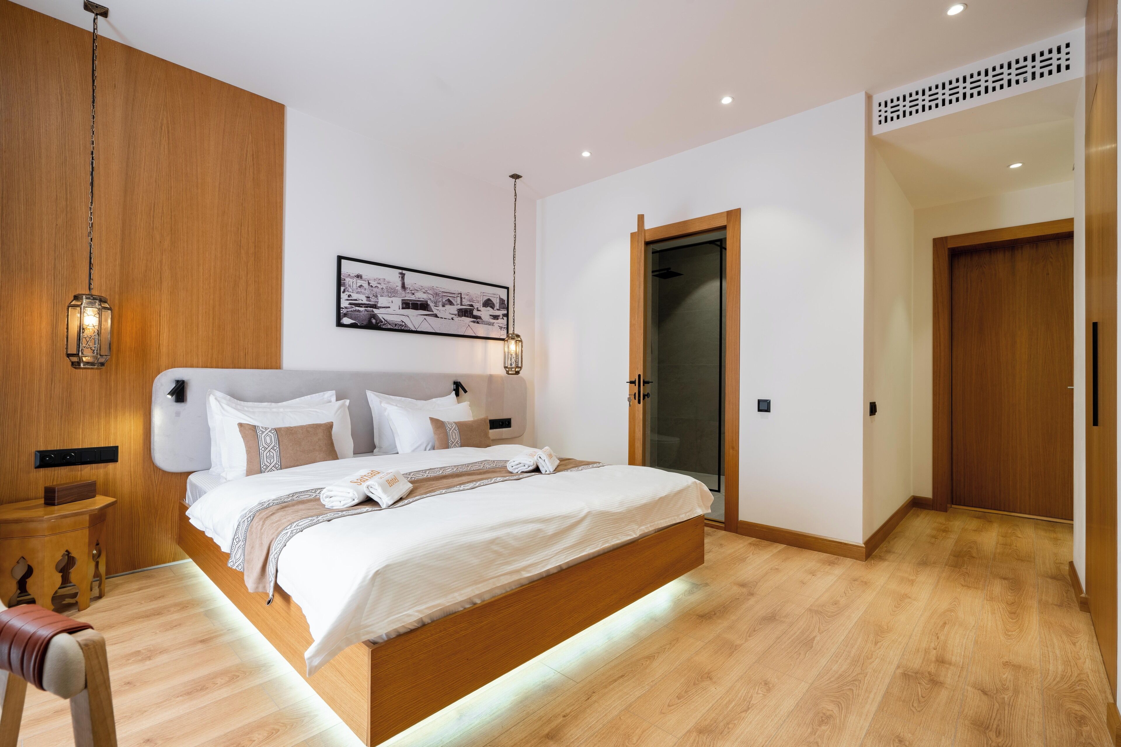 Una habitación de hotel moderna con una cama grande, suelo de madera, una mesita de noche, una silla e imagen enmarcada en la pared.