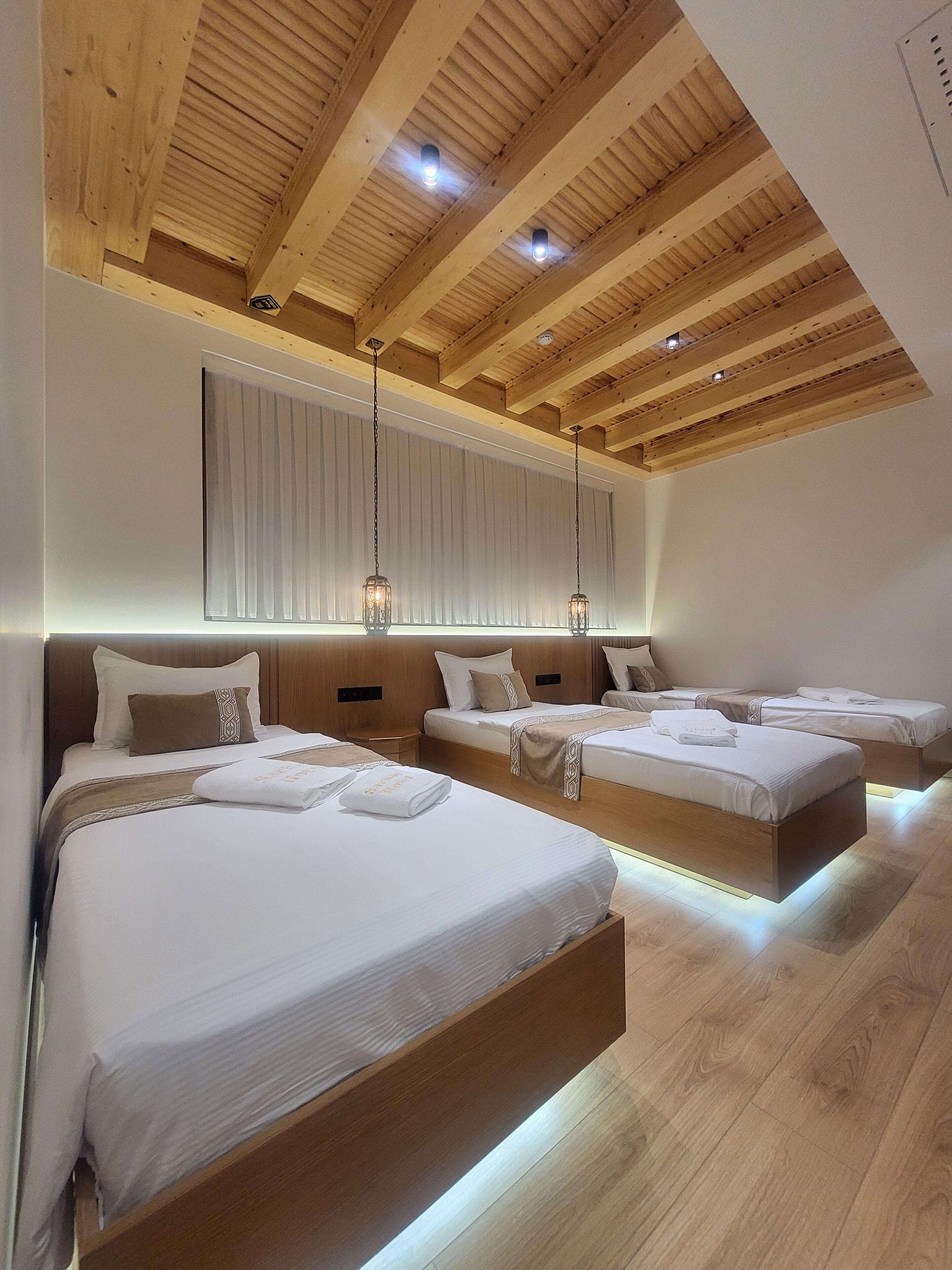 Ein modernes Hotelzimmer mit Holzbalkendecke, drei nebeneinander stehende Betten und Pendelleuchten.