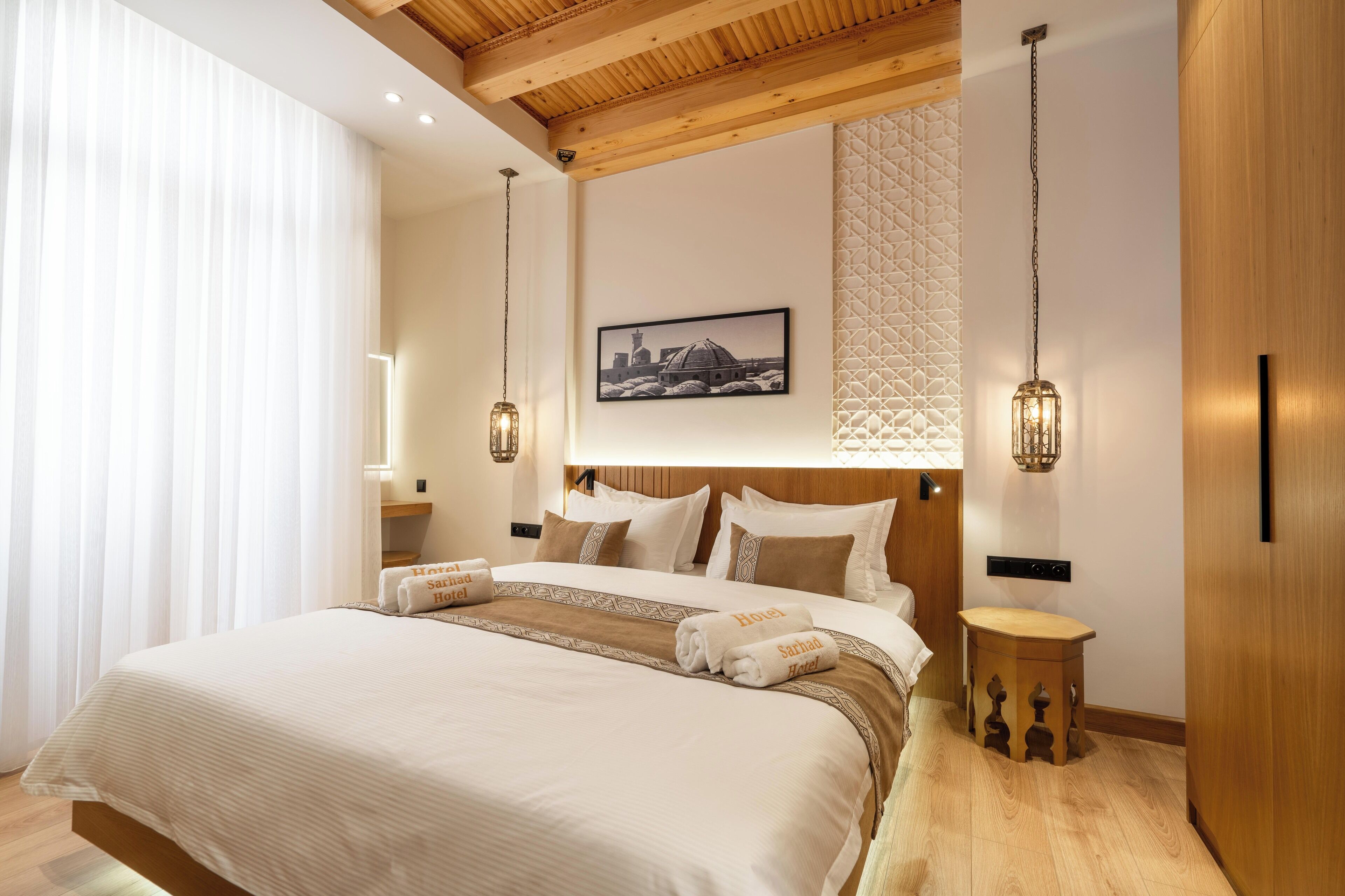Une chambre d’hôtel avec un grand lit, des poutres en bois au plafond, un tableau représentant un paysage urbain encadré et deux lampes fixées au mur.