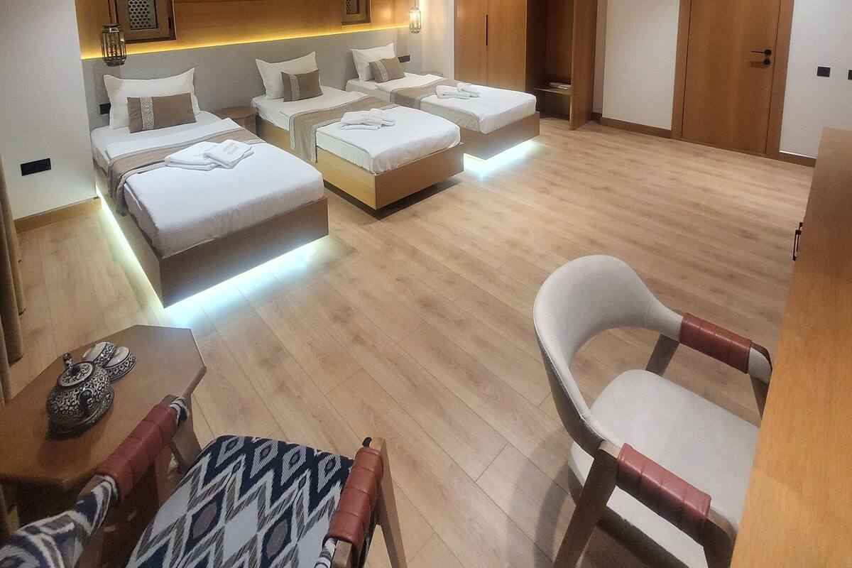 A modern hotel room with a central seating area, multiple beds, a wooden wardrobe, and a small table with decorative items.