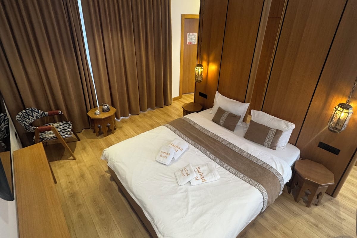 A hotel room with a large bed, wooden panel walls, a small side table, a chair, and a bedside lamp.