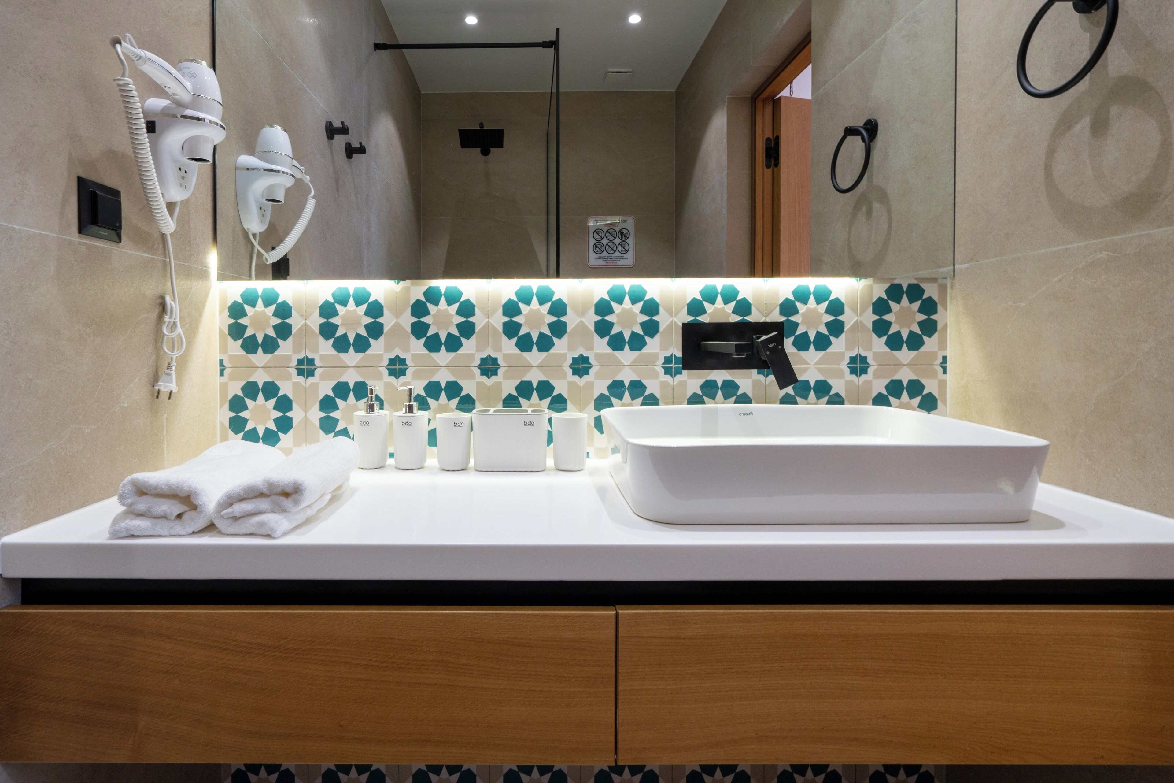 A modern bathroom with a white sink, towel rack, and hair dryers on the wall.
