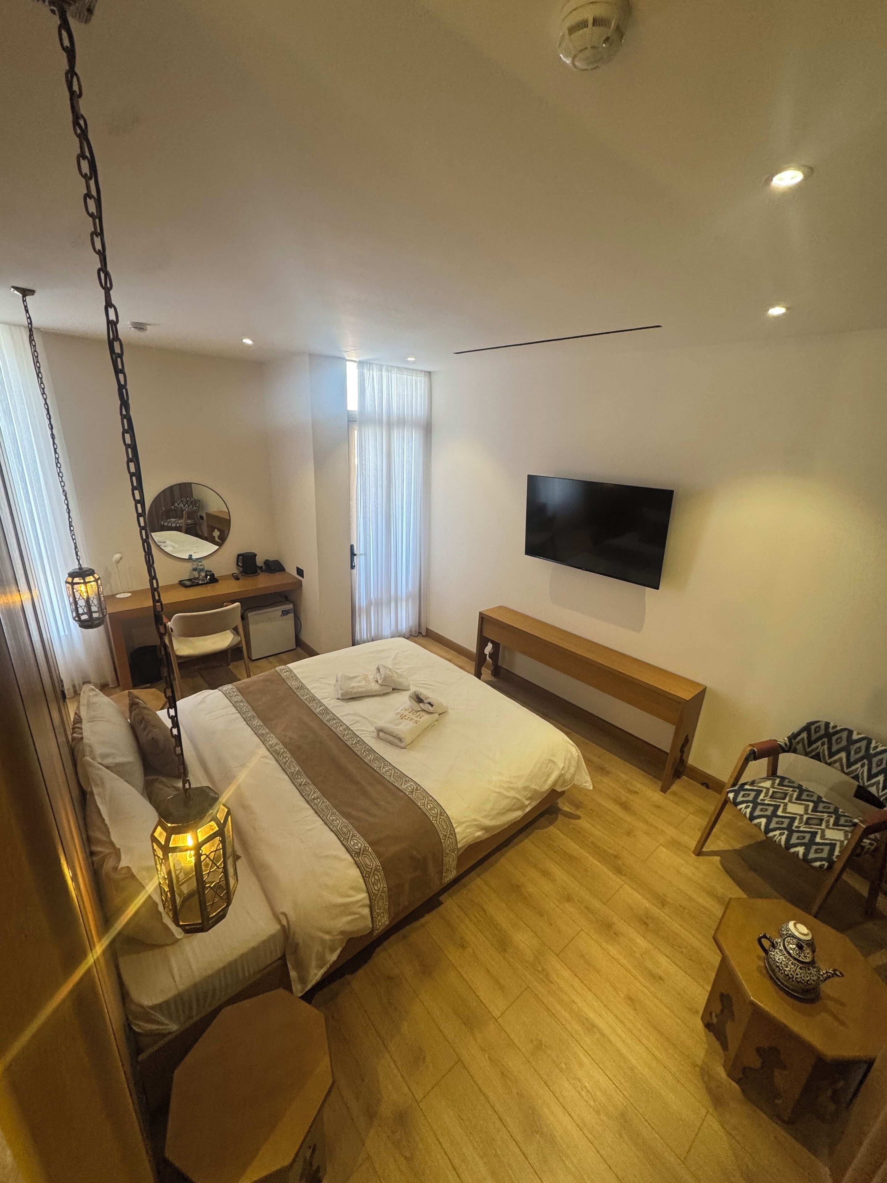 A hotel room with a bed, bedside tables, a desk, a chair, a TV, and a patterned armchair.