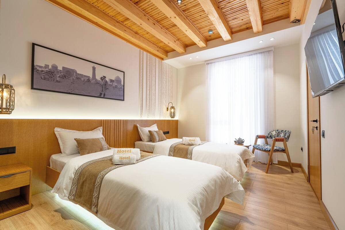 A hotel room with two beds, wooden ceiling beams, a mounted TV, and a cityscape painting on the wall.