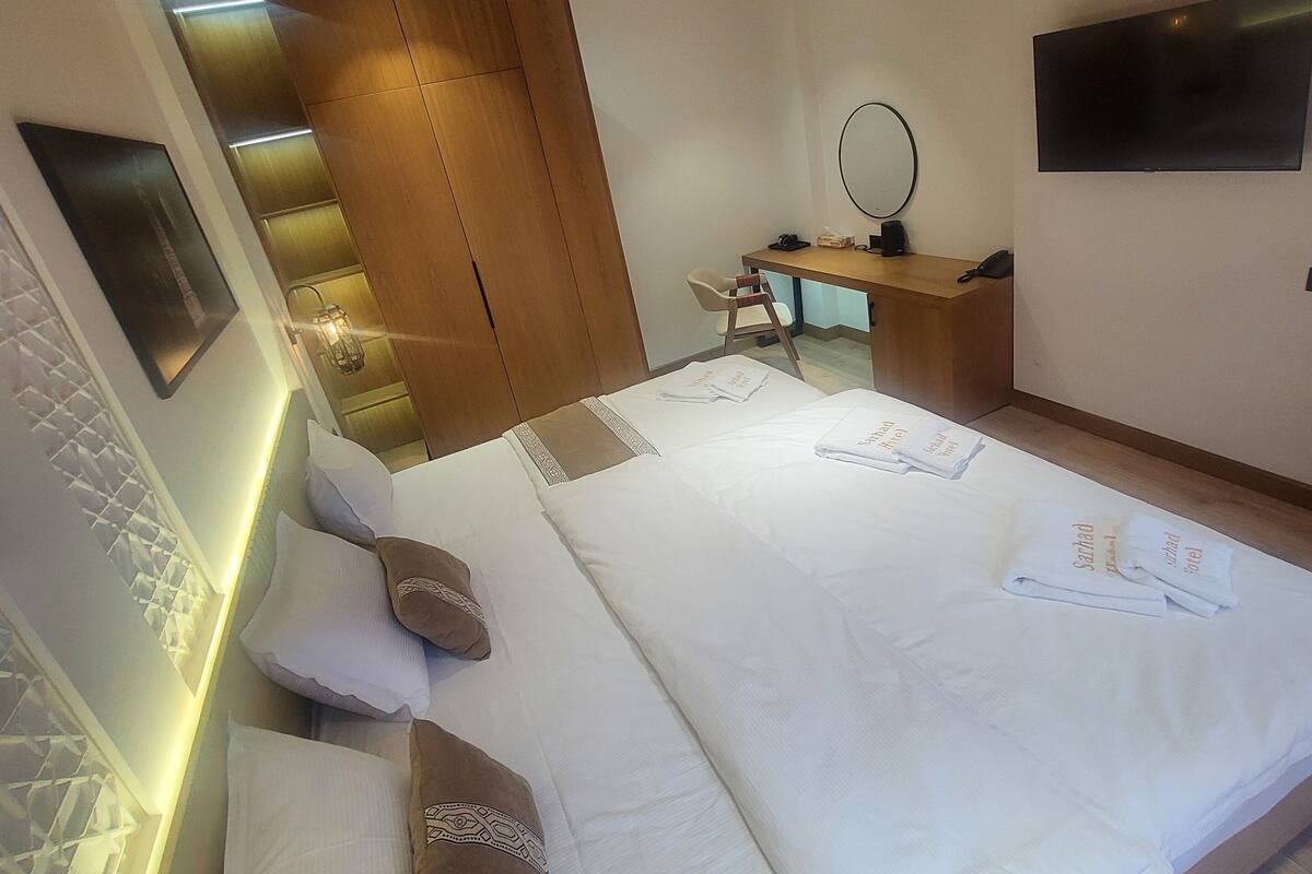A modern hotel room with a large bed, wooden ceiling beams, a desk with a mirror, and a flat-screen TV.