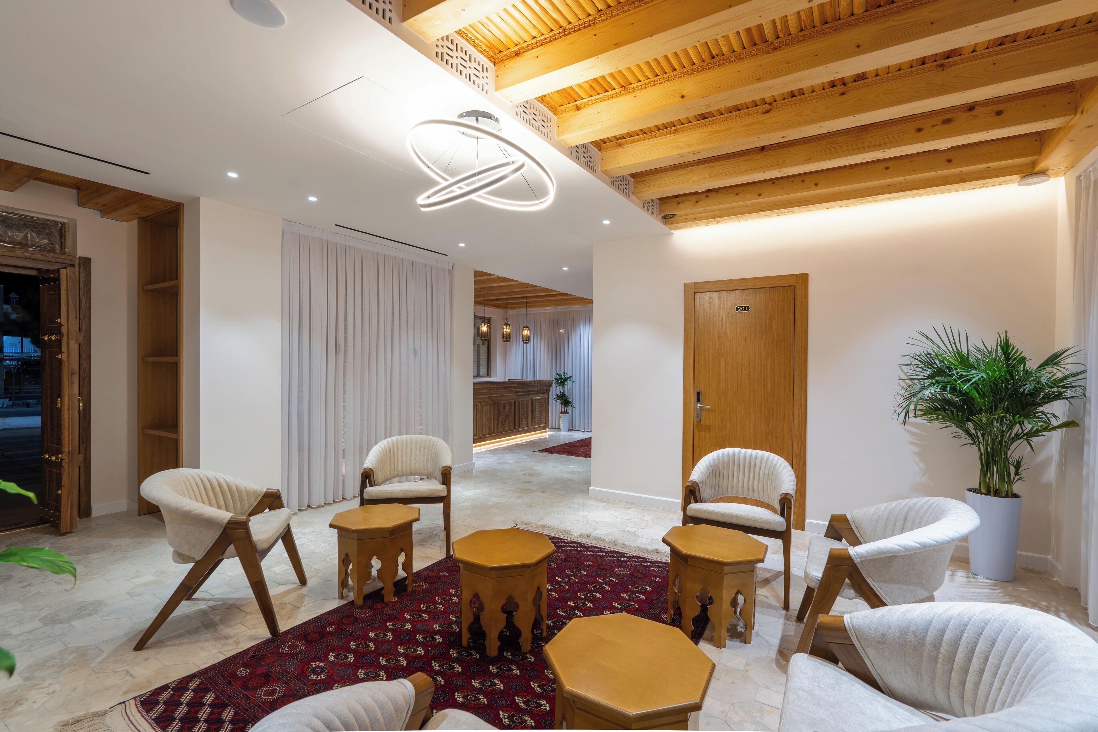A modern interior with wooden beams, white walls, and a red patterned rug. There are several white chairs and a wooden table.