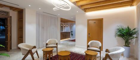 A modern interior with wooden beams, white walls, and a red patterned rug. There are several white chairs and a wooden table.