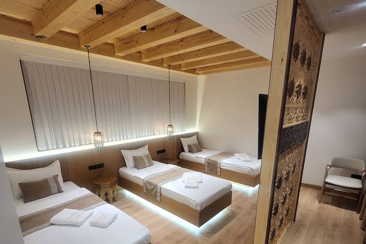 A modern hotel room with three beds, wooden ceiling beams, and a large window with blinds.