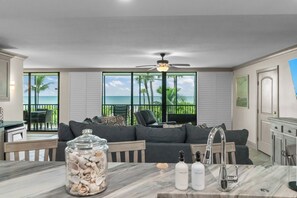 A modern living room with a view of the ocean, a ceiling fan, a glass jar of seashells, and a bar area with bottles.