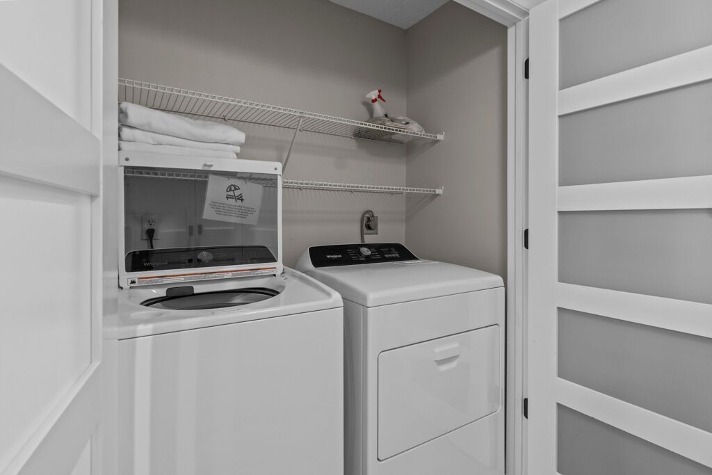 A laundry area with a front-loading washer and dryer, a shelf with folded towels, and a small shelf with a spray bottle.