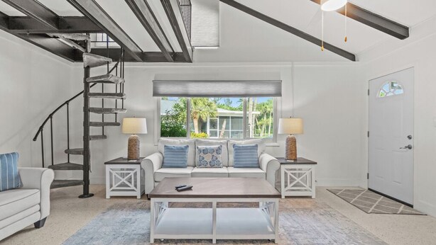 A spacious living room with a white sofa, a coffee table, and a window with a view of greenery. A spiral staircase is visible to the left.