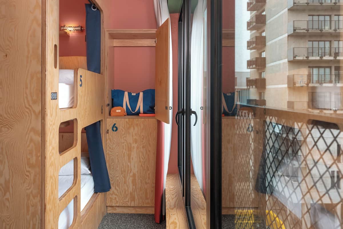 Une chambre avec des lits superposés, un petit coin salon avec un sac, et une vue sur un immeuble à travers une porte vitrée.