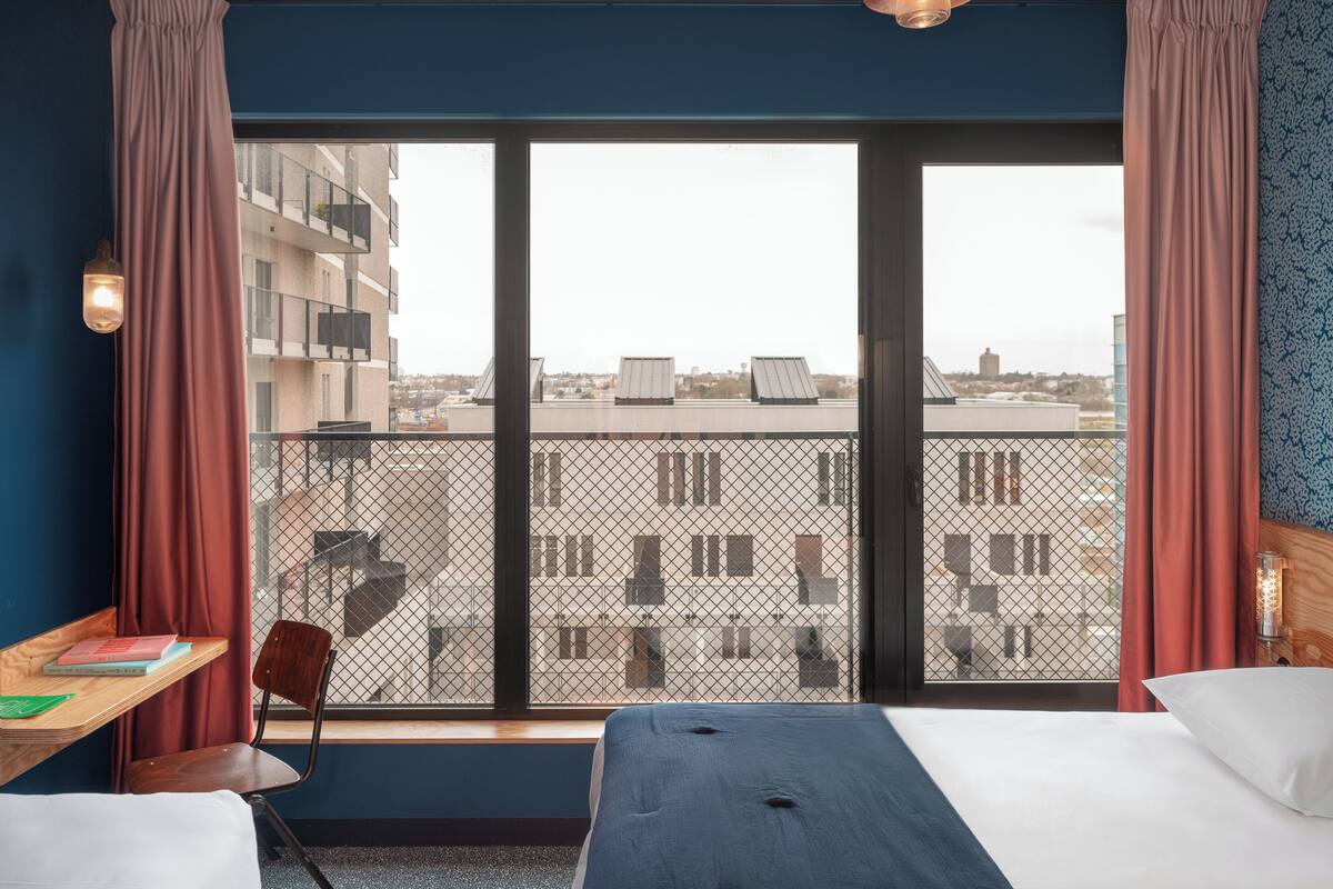 Une chambre d’hôtel avec une grande fenêtre, un lit, un bureau avec une chaise et une petite table.