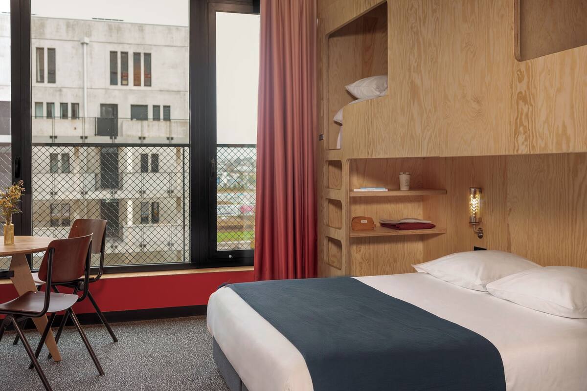 Une chambre d’hôtel comprenant un lit, un bureau avec des chaises, une fenêtre donnant sur des immeubles et une étagère en bois avec divers objets.