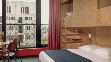 Habitación de hotel con cama, escritorio con sillas, ventana con vista a edificios y estante de madera con objetos.