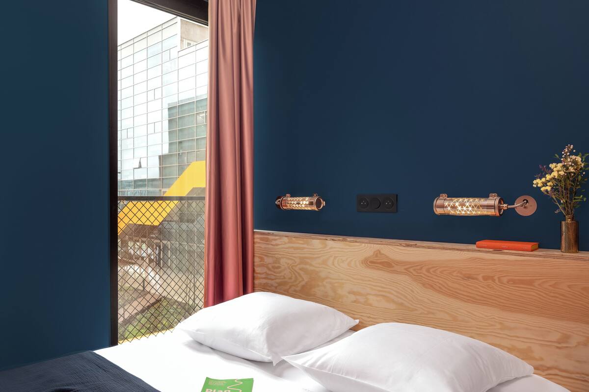 Une chambre d’hôtel avec un lit, une fenêtre donnant sur des immeubles, un rideau et un luminaire fixé au mur.