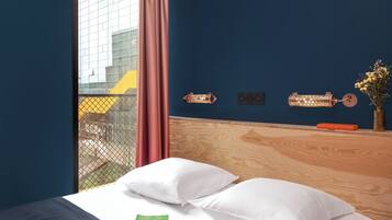 Habitación de hotel con cama, ventana con vista a edificios, cortina y lámpara fijada a la pared.