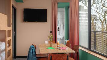 Una stanza con un letto, una TV fissata al muro, un tavolo con sedie, una finestra con tende e vista sull'esterno.
