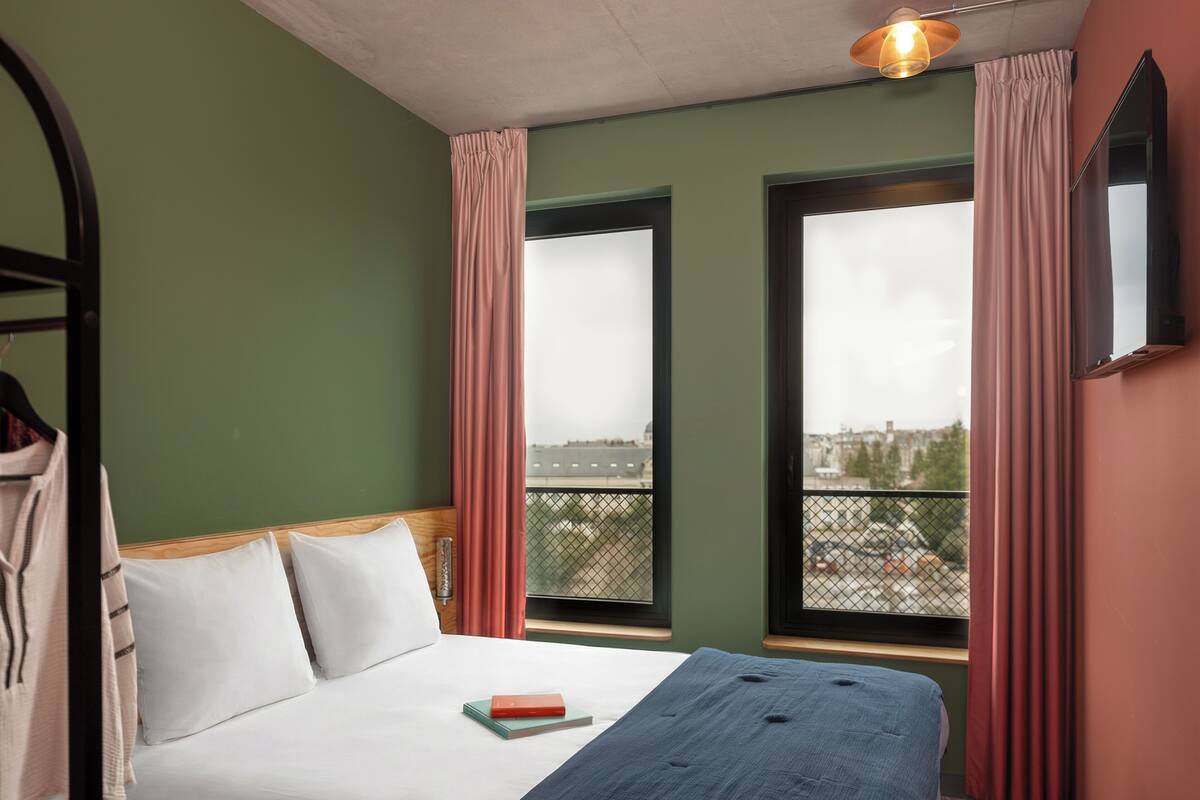 Une chambre d’hôtel avec un lit, deux fenêtres avec des rideaux, une vue sur la ville et un luminaire suspendu.