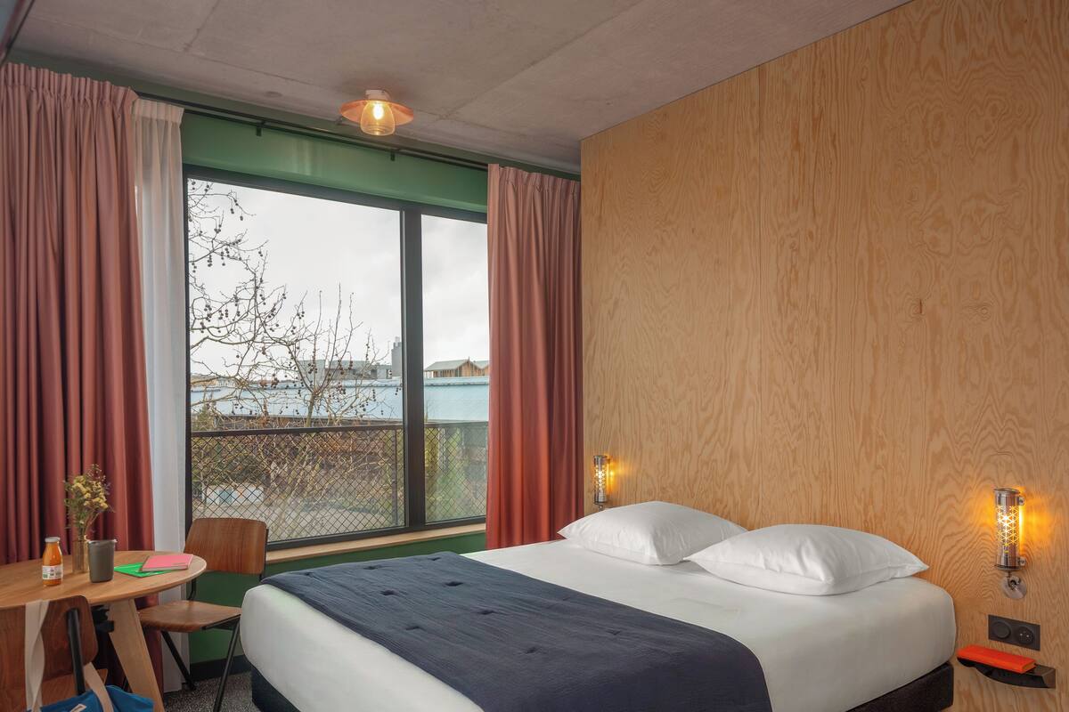 Une chambre d’hôtel avec un lit, un bureau et une chaise, une fenêtre avec des rideaux et une vue sur les bâtiments environnants.