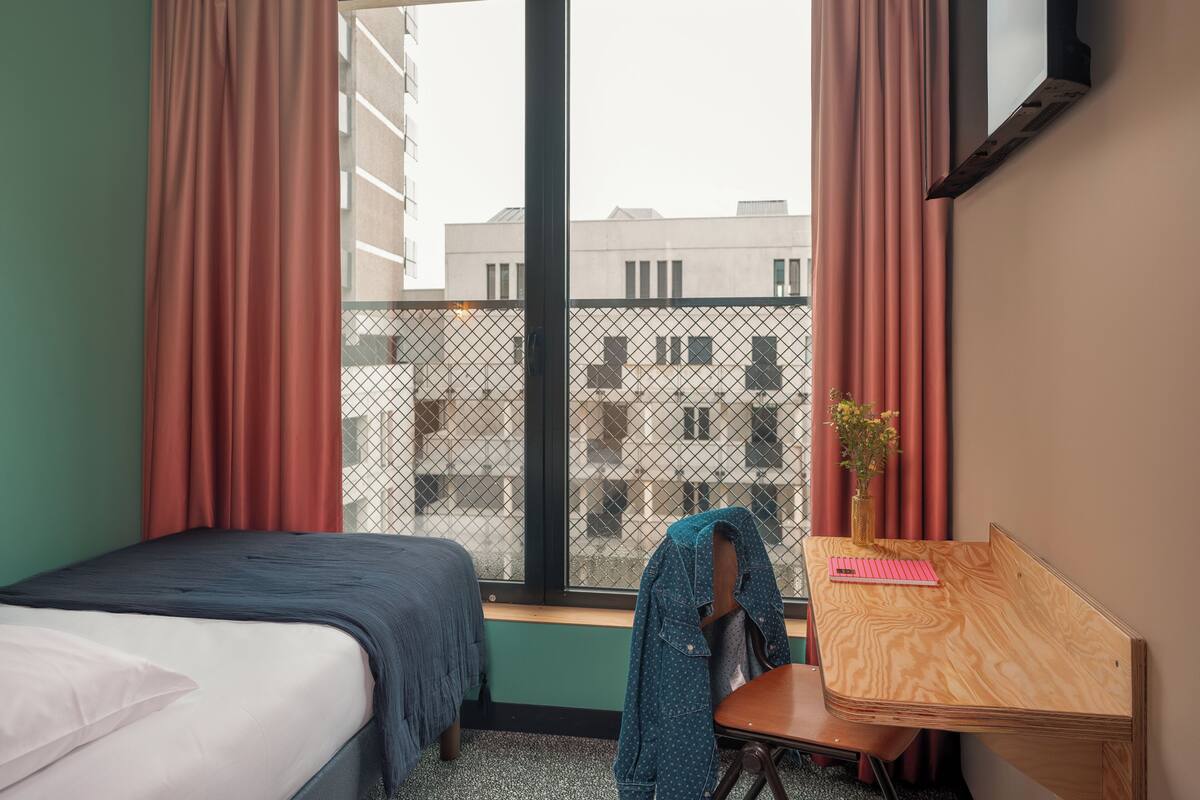 Une chambre à coucher comprenant un lit, un bureau, une chaise, une fenêtre avec des rideaux et une vue sur les bâtiments environnants.