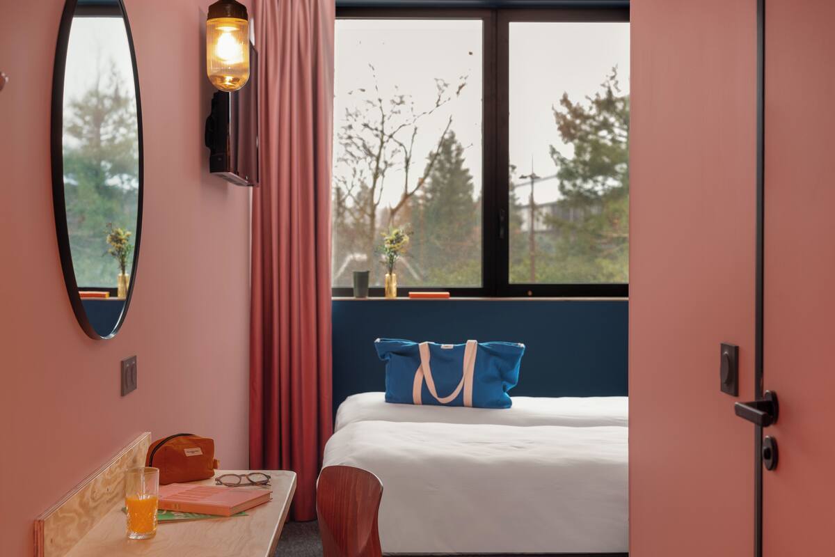 Une chambre avec un lit, un bureau sur lequel se trouve un verre de jus d’orange, un miroir, une fenêtre donnant sur des arbres et un luminaire suspendu.
