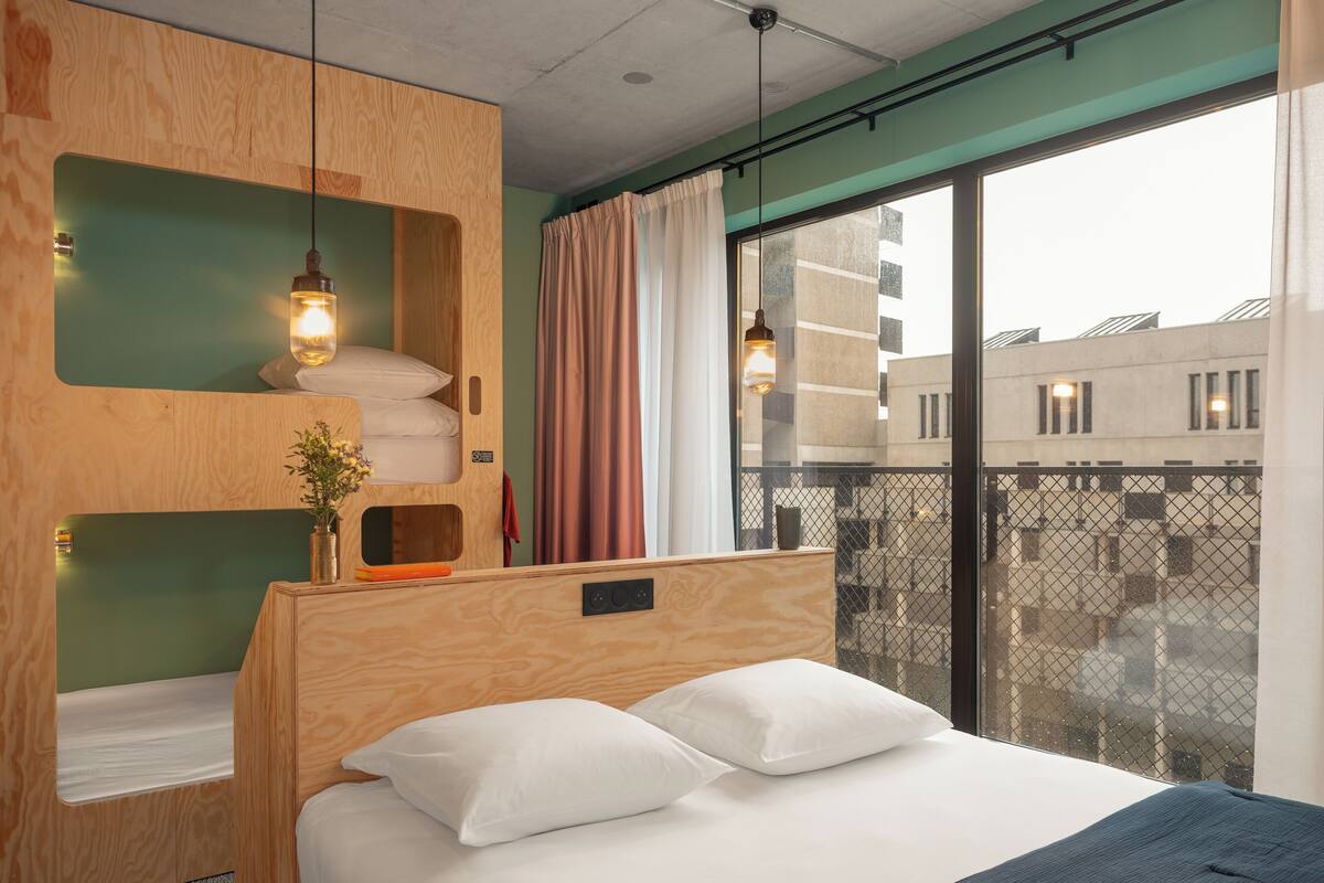 Une chambre d’hôtel moderne, dotée d’un lit, de meubles de rangement en bois, d’une petite table et offrant une vue sur la ville grâce à de grandes fenêtres.
