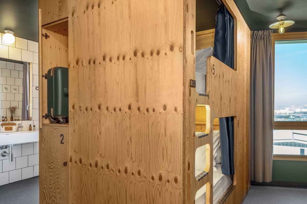 Ein kompakter Raum mit Holztür, auf der '2' steht, einer kleinen Küchenzeile, einem Bett mit Blick und einem Vorhangfenster.