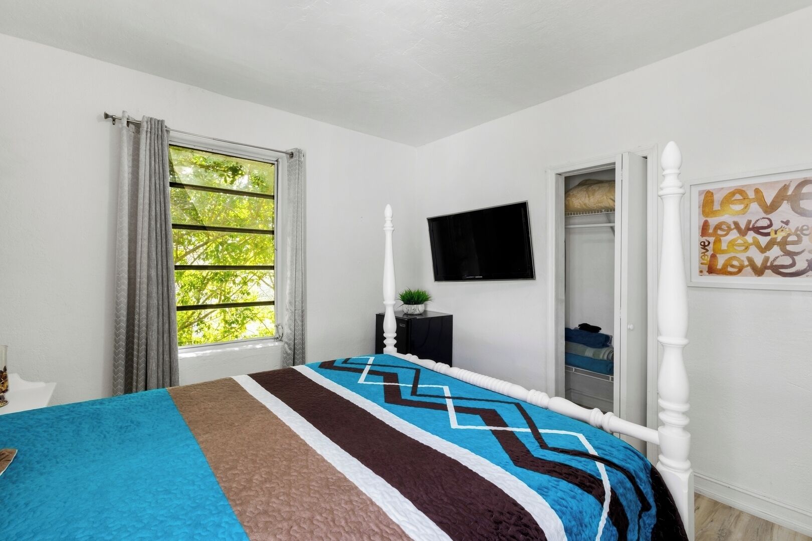 A bedroom with a bed, a TV, a window with curtains, a wardrobe, and a 'Love' artwork.